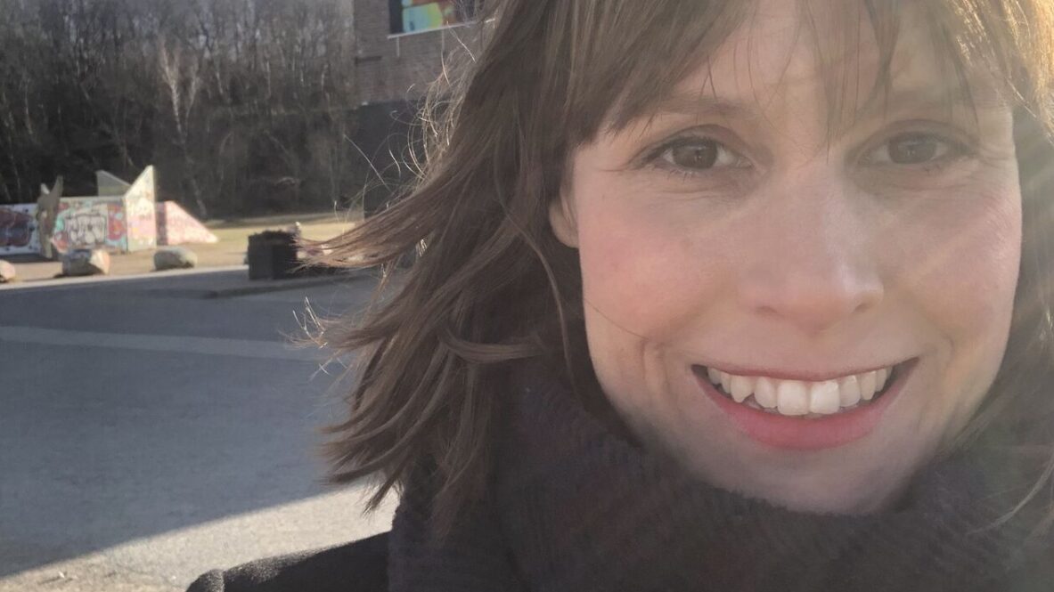 Jenny is wearing a dark coat and a thick scarf standing outdoors in bright sunlight. Behind her is a brick building with a large colorful vertical banner that reads 'BASIM MAGDY' along with exhibition details. In the background, there are leafless trees, a clear blue sky with some clouds, and graffiti on a low structure.
