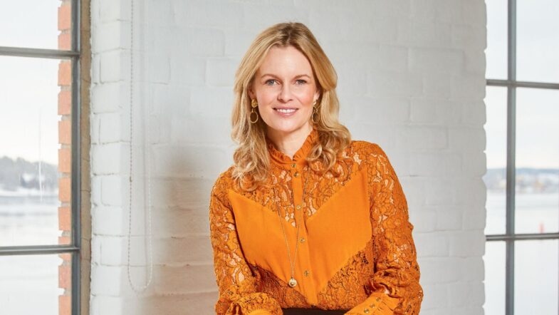Linda is seated indoors against a white brick wall with large windows showing an outdoor view. She is wearing an orange lace blouse with decorative buttons and a long necklace, paired with a black skirt and dark tights. Hands are resting on the lap, and the lighting is bright and natural.