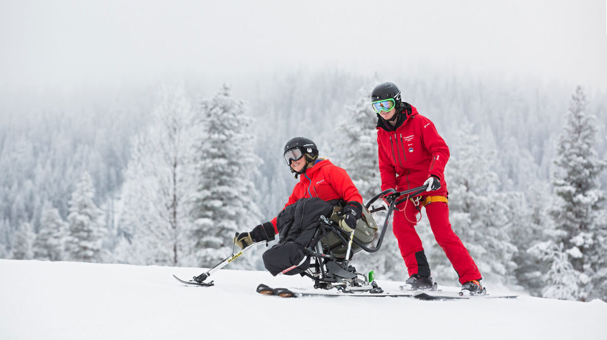 En person åker sitski med stöd av en skidlärare, båda ler. Snötäckta träd i bakgrunden