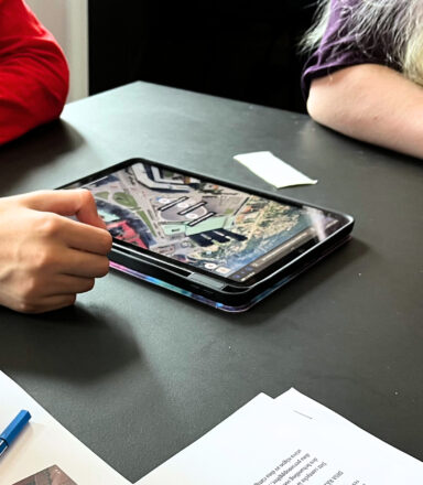 Två personer sitter vid ett bord med en surfplatta som visar en stadskarta. Framför dem ligger papper och en penna.