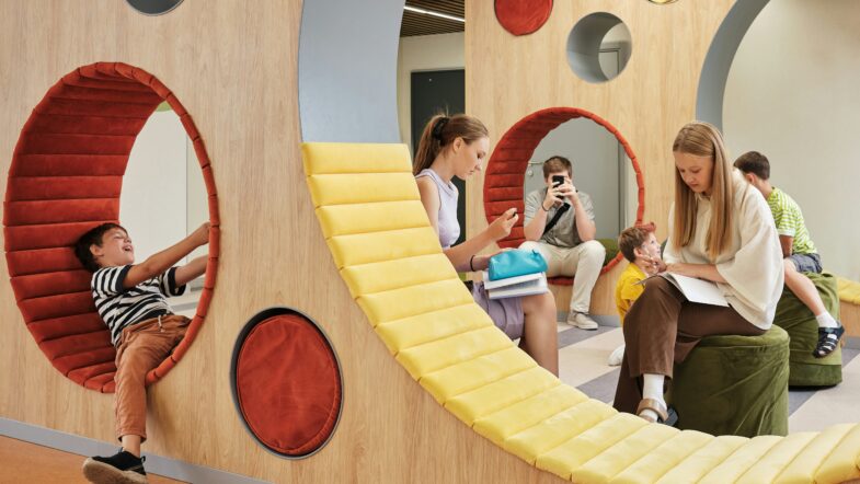 En vägg med stora och små cirkulära öppningar klädda med röda och gula dynor. Ett barn sitter i en öppning, och flera personer sitter på gröna sittpuffar framför väggen.