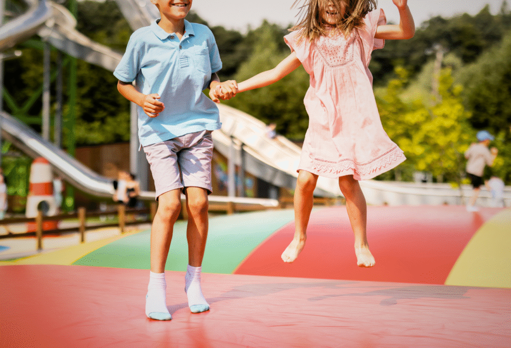 Två barn hoppar och håller varandra i handen på en stor färgglad hoppkudde utomhus. I bakgrunden syns rutschkanor och gröna träd.