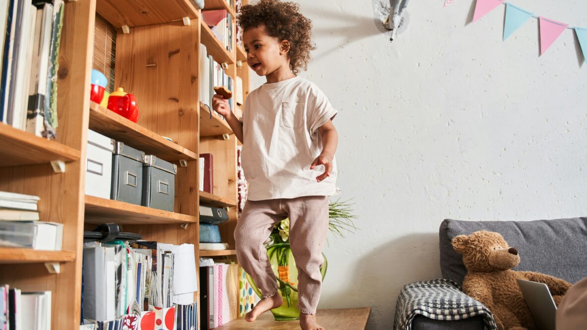 Ett barn står på en låg träbänk framför en stor bokhylla av trä. Barnet bär en ljus kortärmad tröja och mönstrade byxor i brunt och beige. Barnet sträcker sig mot en av hyllorna som innehåller böcker, pärmar, lådor och några färgglada föremål, bland annat en röd leksak. På bänken ligger en grön leksak. Till höger i bilden syns en soffa med en gråvit rutmönstrad filt och en stor brun nalle som lutar mot armstödet. På väggen bakom soffan hänger en rad färgglada vimplar i rosa, blått och grönt. Ljuset i rummet är mjukt och naturligt.