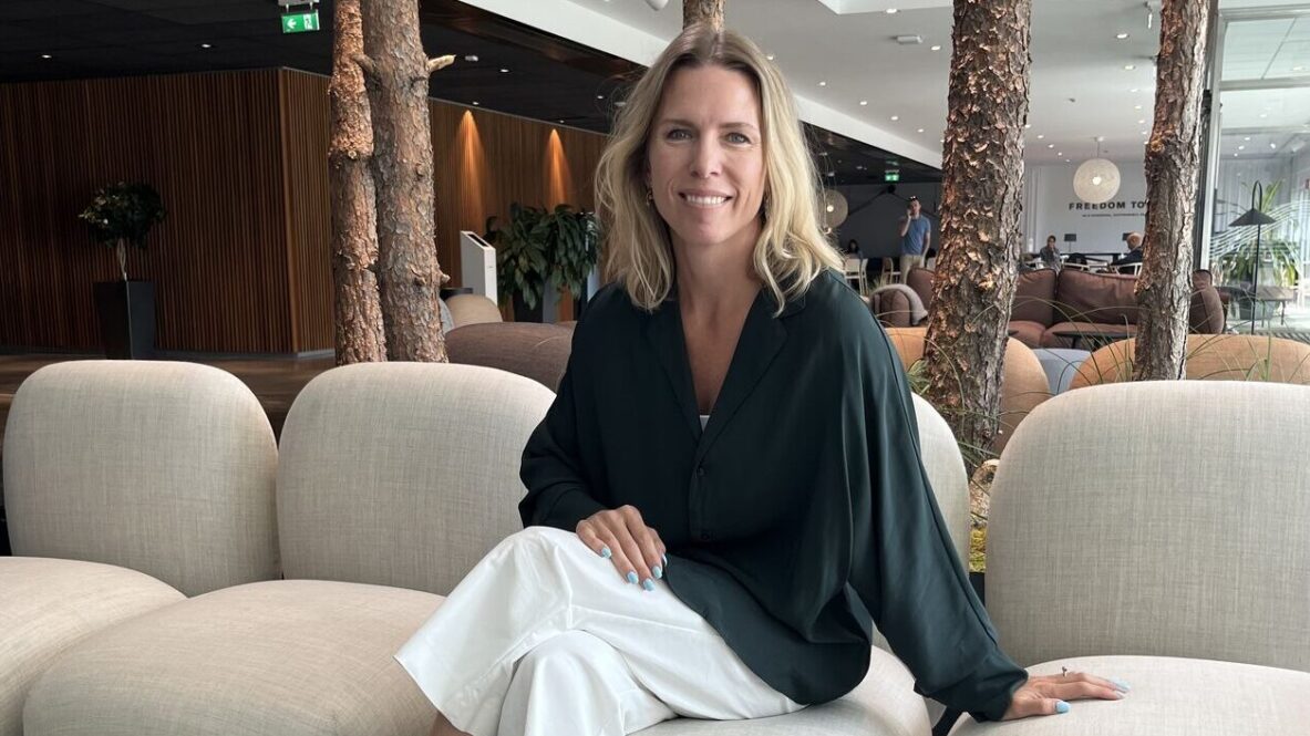 Emelie is seated on a light beige sofa in a modern indoor lounge area. She is wearing a dark green long-sleeved top, white pants, and black ankle boots. Behind the sofa are several vertical tree trunks integrated into the design, and in the background there are wooden wall panels, plants, and additional seating.