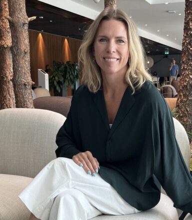 Emelie is seated on a light beige sofa in a modern indoor lounge area. She is wearing a dark green long-sleeved top, white pants, and black ankle boots. Behind the sofa are several vertical tree trunks integrated into the design, and in the background there are wooden wall panels, plants, and additional seating.