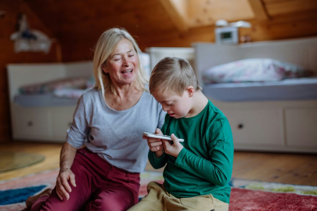 An adult and a child sit on the floor in a bedroom, the child is holding and looking closely at a smartphone while the adult sits beside them.