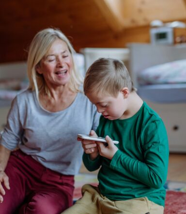 An adult and a child sit on the floor in a bedroom, the child is holding and looking closely at a smartphone while the adult sits beside them.