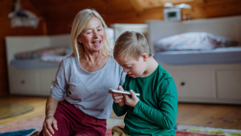 An adult and a child sit on the floor in a bedroom, the child is holding and looking closely at a smartphone while the adult sits beside them.