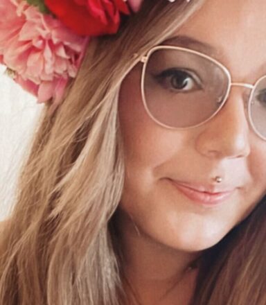 Sara-Karolina is wearing a floral-patterned shirt and a necklace with a round pendant is adorned with a large flower crown made of pink and red blossoms with green leaves. The background appears softly lit indoors.