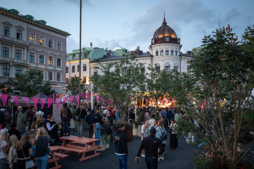 An outdoor festival scene with a large crowd gathered among trees, pink flags, and market tents in a city square. Historic buildings with decorative facades and a domed rooftop surround the lively evening event.