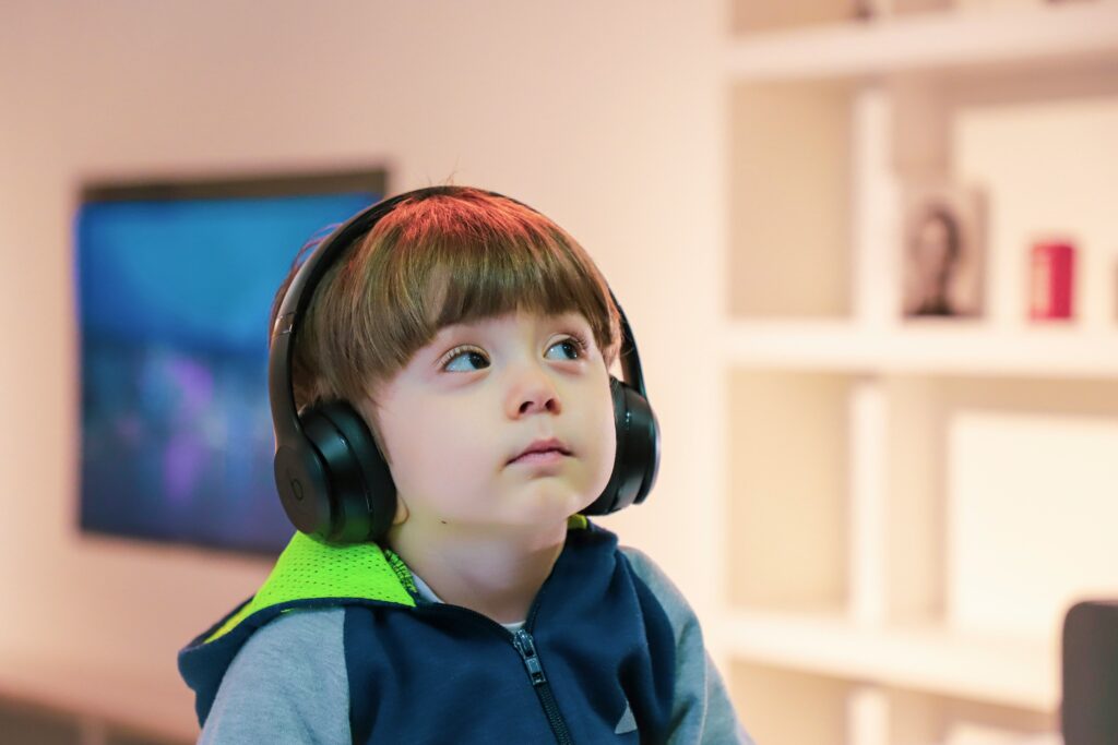 A child wearing black over‑ear headphones sits indoors in front of a television screen, dressed in a blue hoodie with a bright green hood.