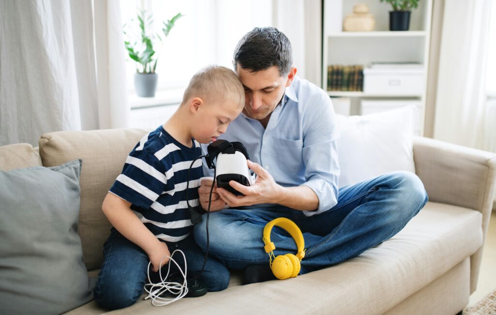 An adult and a child sit together on a sofa while looking at a virtual reality headset that the adult is holding. A pair of yellow headphones and a coiled cable lie on the sofa beside them.