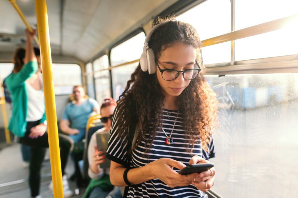 A person with long curly hair wearing white headphones stands inside a bus using a smartphone, while other passengers sit and stand nearby along the aisle.