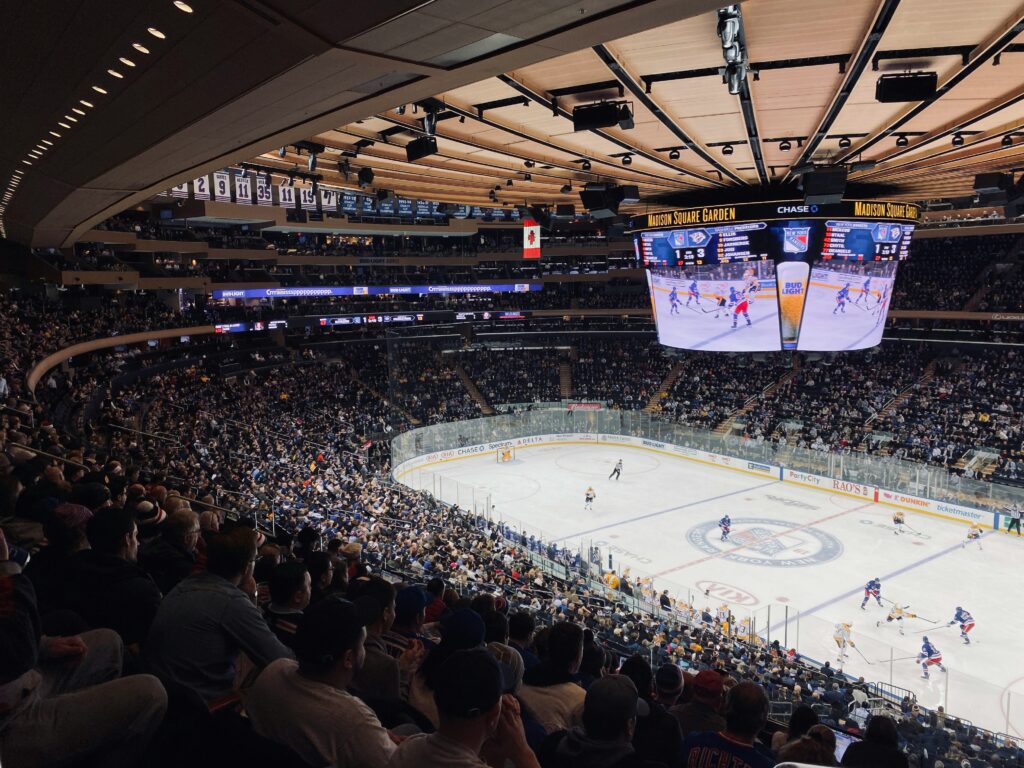 A large indoor arena filled with spectators watching an ice hockey game. Players skate across the rink below, and a giant overhead screen displays the action above the crowd.